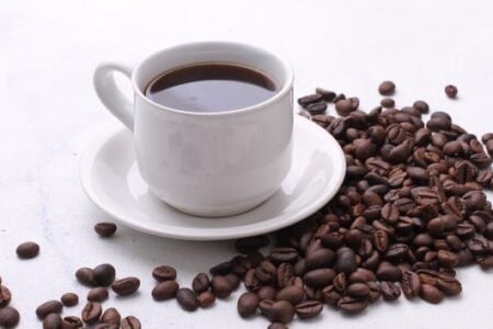 A white ceramic cup full of black coffee on a saucer, surrounded by scattered dark roasted coffee beans.
