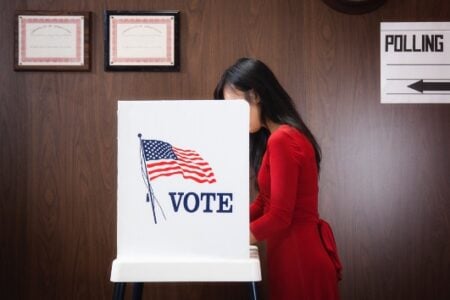 Woman in a red dress voting at a booth with an American flag and "VOTE" sign inside a polling place.