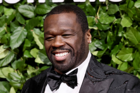 Cedric the Entertainer smiles while wearing a black tuxedo and bow tie against a green leaf wall.