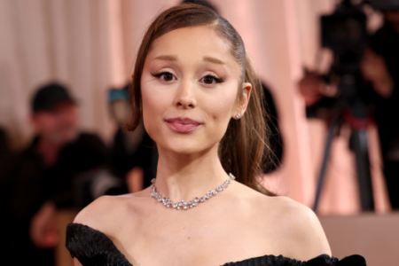 Ariana Grande poses on a red carpet in a black off-the-shoulder dress and diamond necklace, looking at the camera.