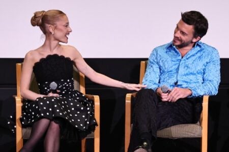 Ariana Grande and Ethan Slater share a laugh, sitting on stage, both holding microphones, with Grande touching Slater's arm.