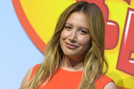 Ashley Tisdale smiles slightly, wearing an orange top, against a colorful blue, yellow, and red backdrop.