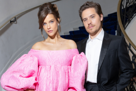 Barbara Palvin in an off-shoulder pink dress and Dylan Sprouse in a black suit stand by a curved staircase with blue carpet.