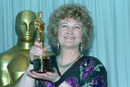 Smiling woman with curly blonde hair holds an Oscar, a large golden Oscar statue behind her against a blue curtain.