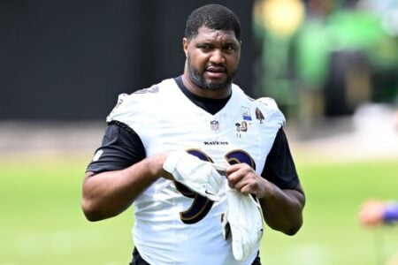 Calais Campbell, wearing a white Baltimore Ravens #99 jersey, adjusts his white gloves on a sunny practice field.