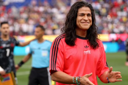Former Mexican goalkeeper Jorge Campos smiles and gestures in a red shirt on a soccer field.