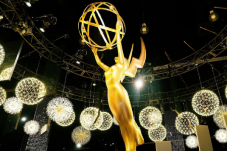 Gold Emmy statue standing illuminated amidst glowing spherical lights and stage trussing.