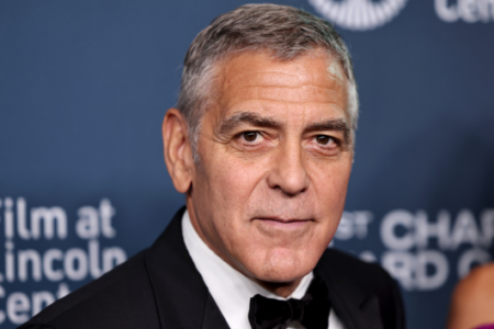 George Clooney in a black tuxedo and bow tie, looking directly at the viewer, stands before a blue 'Film at Lincoln Center' sign.