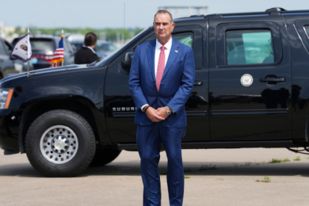 Man in blue suit stands by a black armored SUV with US and Vice Presidential flags on a paved surface.