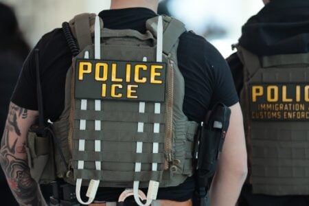 Two ICE agents, viewed from behind, wearing tactical vests marked "POLICE ICE" and "IMMIGRATION AND CUSTOMS ENFORCEMENT.