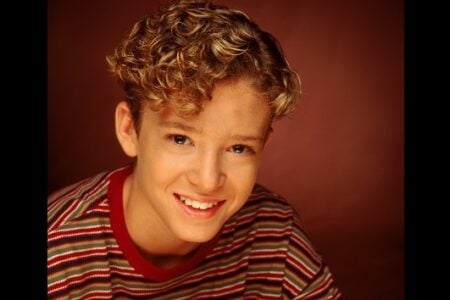 Young Justin Timberlake with curly blonde hair and a striped shirt smiles broadly against a reddish-brown background.