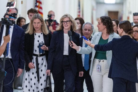 Senator Kyrsten Sinema walks down a hallway, speaking into a reporter's microphone, surrounded by staff and press.