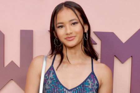 Actress Miya Cech smiles, wearing a blue patterned top, against a pink wall with large purple letter shapes.