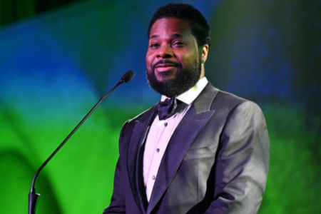 Bearded man in a tuxedo smiles at a microphone, against green and blue stage lighting.