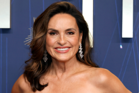 Mariska Hargitay smiling broadly, wearing a strapless gown and diamond earrings, against a dark blue event backdrop.