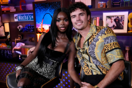 A smiling Black woman and White man sit holding hands on a vibrant talk show set filled with eclectic decorations.
