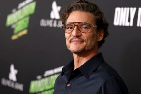 Pedro Pascal, with a mustache and glasses, smiles gently at a premiere against a dark background with blurred logos.