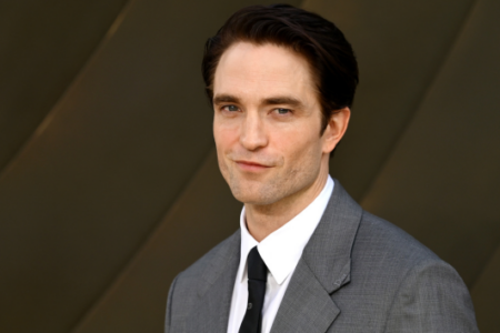 Robert Pattinson in a grey suit and black tie, looking at the camera with a slight smile against a dark architectural background.