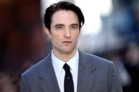 Actor Robert Pattinson in a grey suit and tie looks directly at the camera outdoors.
