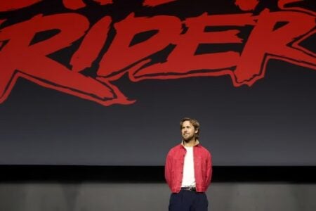 Ryan Gosling stands on a stage in a red jacket, looking right, with "RIDER" in red text on the dark screen behind him.