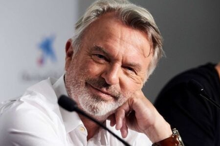 Actor Sam Neill smiles softly, resting his chin on his hand, at a press conference with a microphone in front of him.