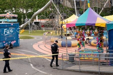 Police officers investigate a carnival area cordoned off with yellow tape, featuring game tents and a "Fun" stand.