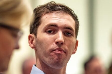 Young man in a blue shirt with a serious expression looks at the camera amidst a blurred crowd indoors.