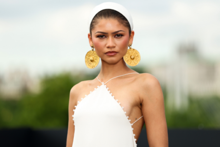 Zendaya, in a white headband, large gold earrings, and white halter dress, poses outdoors against a blurred background.