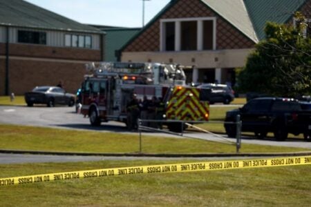 POLICE LINE DO NOT CROSS" tape in foreground, with a fire truck and vehicles outside a building with a green roof.