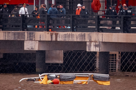 NYPD officers and firefighters on a pier overlook a floating yellow rescue stretcher and gear in the water below.