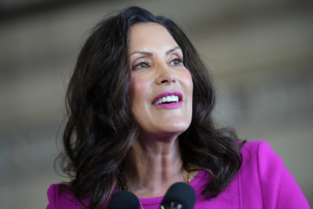 Governor Gretchen Whitmer speaks into two microphones, wearing a fuchsia jacket, indoors.