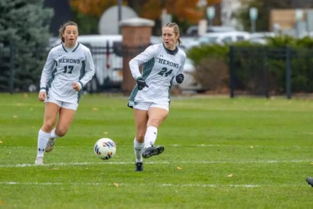 Herons soccer player #24 dribbles a ball down a grassy field as teammate #17 runs behind her.