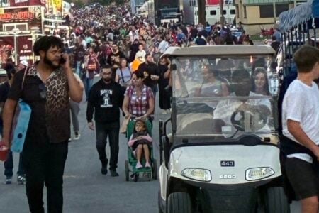 A large crowd walks through an outdoor fairground at dusk, past vendor stalls and a golf cart under a pink sky.