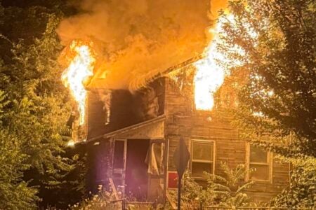 Two-story house fully ablaze with large flames and smoke erupting from the roof and windows at night, seen from the street.