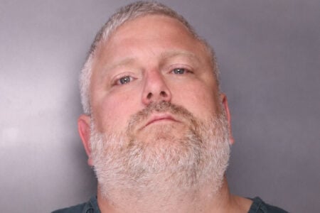 Mugshot of a man with graying hair and beard, wearing a dark shirt, looking directly at the camera against a plain background.