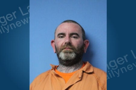 Bearded man with neck tattoos in an orange jumpsuit looking forward for a mugshot against a plain blue background.