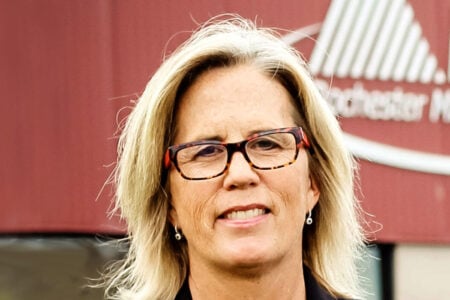 Blond woman with glasses and black blazer smiling in front of a red Rochester Midland building.