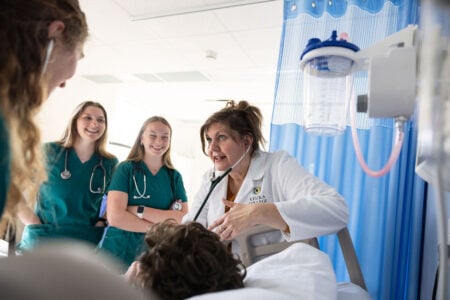 Keuka College nursing instructor uses a stethoscope on a patient, teaching three smiling students in a medical simulation.