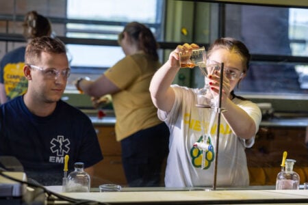 A female student in safety goggles pours a liquid from a beaker into a separatory funnel in a science lab, while a male student watches.