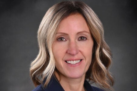 Smiling woman with wavy blonde hair in a dark blazer against a gray background.