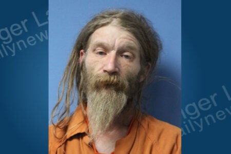 Mugshot of a man with long, light brown hair and a long, scraggly beard, wearing an orange jumpsuit against a blue background.