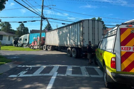 A damaged white semi-truck and other rigs are stuck at a residential intersection, attended by emergency services.