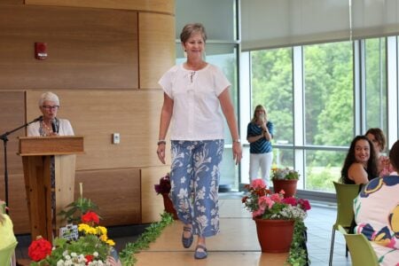 Smiling woman in white top & blue floral pants walks an indoor runway past a podium speaker.