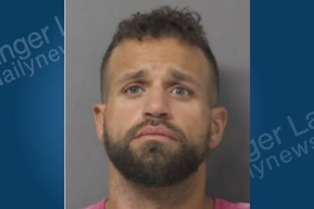 A man with a dark beard, short hair, and light eyes looks directly at the camera in a booking photo.