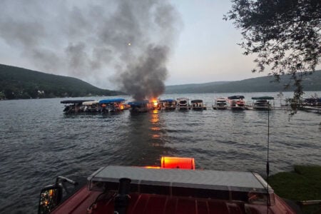 Boat ablaze at Finger Lake dock, thick black smoke rising against evening sky, seen from emergency vehicle.