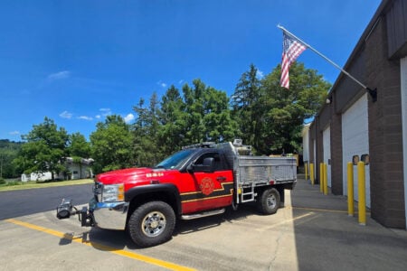 Red wildland fire truck 62-M-54 parked at a fire station on a sunny day with an American flag.
