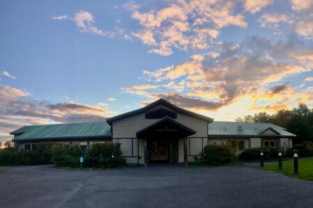 Long, low building with a dark green roof and covered entryway at dawn or dusk, framed by a colorful cloud-filled sky.