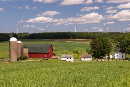 Red barn, silos, and green farm fields under a blue sky, with wind turbines lining a distant treeline.