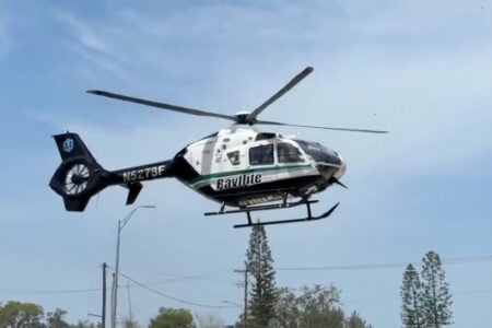 A black, white, and green Bayflite medical helicopter (N5278F) flies low over trees and power lines.
