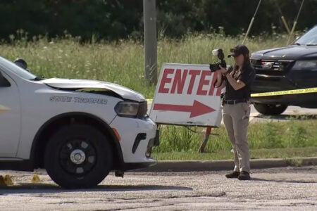 A State Trooper photographs a damaged police vehicle with evidence markers at a roadside incident scene.